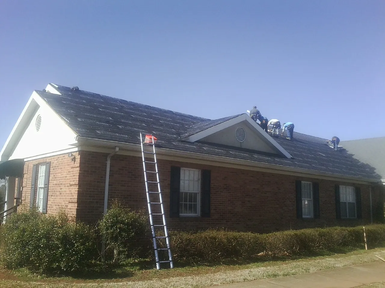 Crew tearing shingles off a brick ranch roof with a ladder set — Metal Roof Repair work by our crew in Richford