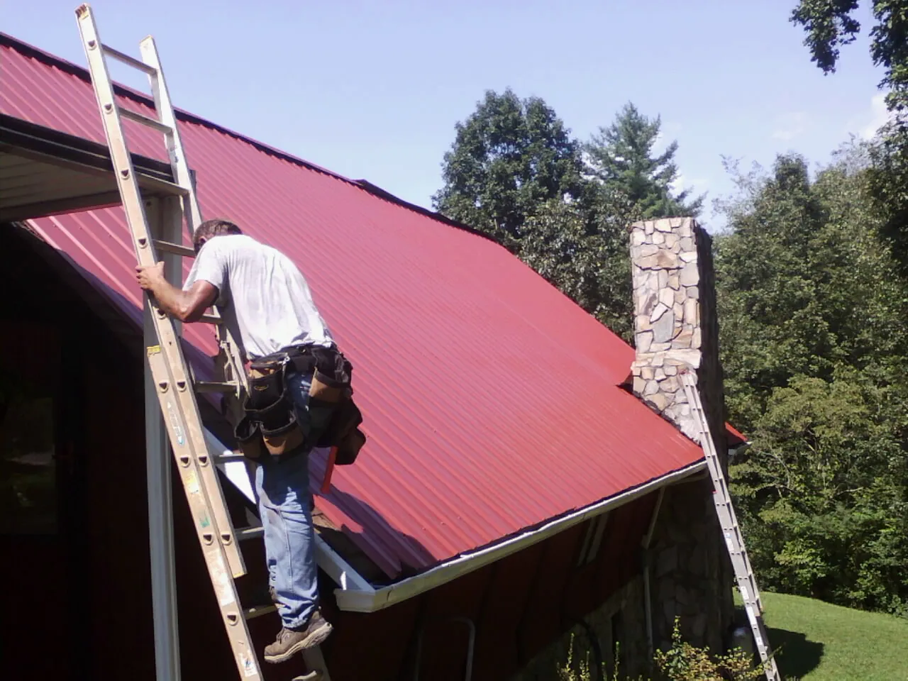 Roofer on a ladder beside a red metal roof and stone chimney — Soffit & Fascia Repair by Roofing Brothers in Richford